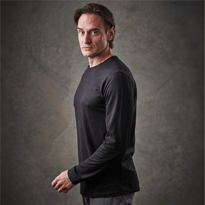 A man wearing a black long sleeve t-shirt, posing against a textured background. The shirt features branding.