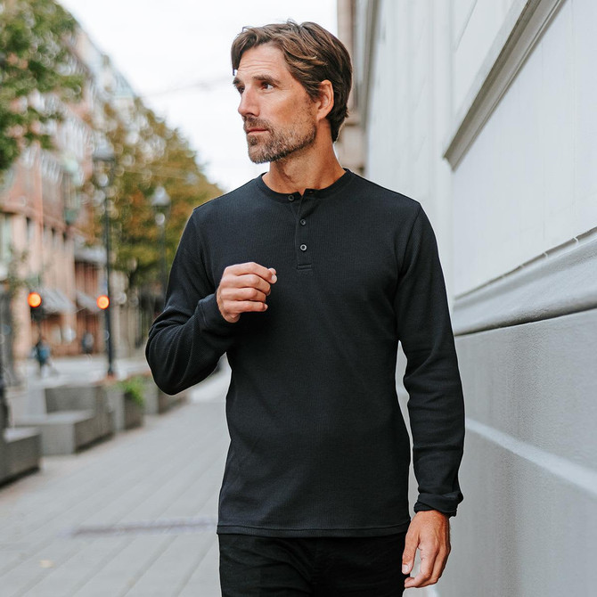 A man walks outdoors wearing a black long-sleeve henley shirt, featuring a three-button placket. The background shows a city street.