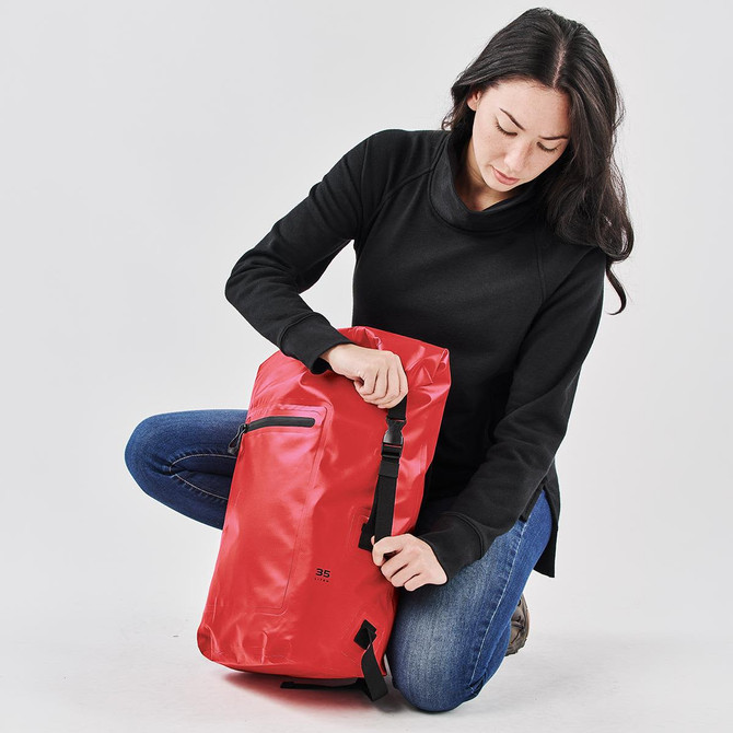 A red backpack with a sleek design, featuring a zippered pocket and adjustable straps, held by a person in black attire.