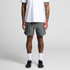 A person stands wearing grey swim shorts, paired with a white t-shirt and black sneakers. The shorts feature a logo.