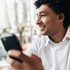 A man with curly hair is smiling while holding a smartphone and wearing black wireless earbuds indoors.
