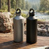 A drink bottle set in grey and black, featuring a carabiner, placed on a wooden surface near a stream.