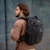 A man with long hair wearing a brown jacket carries a black Swissdigital Neptune Backpack. The backpack has a logo.