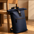 A navy roll-top backpack with a front zip pocket rests against a chair in a warm, indoor setting.