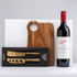 A drink bottle of red wine alongside a wooden cheese board and a gold two-piece cheese knife set, featuring a branded box.