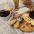 A wooden cheese board with a round cheese, crackers, and a bowl of olives, accompanied by cheese knives.
