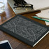 A black recycled cotton cahier notebook with a leaf pattern, placed on a wooden desk among office supplies.