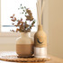 A wooden reed diffuser with light wood finish and three reeds beside a cream and terracotta vase with dried foliage.