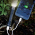 A black power bank resembling a torch is connected to a smartphone on a natural background with greenery.
