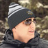 A black and grey beanie with a logo, worn by a person in a winter setting, featuring sunglasses and a jacket.