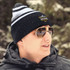A black and grey striped beanie is worn by a person in sunglasses and a padded jacket, surrounded by snow.