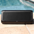 A black outdoor speaker with a mesh front, placed on a stone surface by a pool. It has branding on the side.