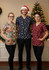 Three models wear festive unisex scrub tops in red, navy, and multicoloured designs, with a Christmas tree in the background.