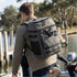 A grey backpack cooler, with multiple compartments, being carried by a person near a waterfront.