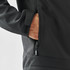 A black softshell jacket with a zippered pocket, featuring a hand reaching inside the pocket.
