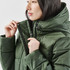A woman in a green puffer jacket adjusts the zipper, showcasing the jacket's high collar and insulation.