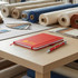 A red notebook and a red pen on a wooden table, surrounded by fabric rolls and colour swatches.