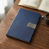 A navy blue A5 notebook with a textured cover and a silver clasp, placed on a wooden table next to a coffee cup.