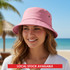 A pink bucket hat sits on a smiling woman's head with a beach backdrop and palm trees. The hat features a logo.