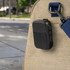 A black Bluetooth speaker attached to a bag, set against an outdoor urban background. It features controls and a logo.