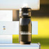 A drink bottle in black with a grip design, featuring a logo and a nozzle cap on a metallic surface.