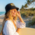A woman wearing a black terry towelling bucket hat with a full-colour patch, smiling on a beach with palm trees.