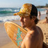 A man stands on the beach wearing a yellow floral trucker cap with a logo, holding a surfboard.