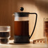 A Bodum Brazil French Press in black with a glass body, sitting next to a glass cup of coffee on a wooden shelf.