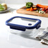 A glass lunch box with a navy blue plastic lid, placed on a kitchen counter next to fresh vegetables and a towel.