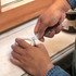 A hand using a silver bottle opener key ring to open a paint tin, resting on a light-coloured surface.