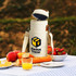 A cream wine cooler bag with a logo, resting on a picnic table surrounded by apples, strawberries, bread, and juice.