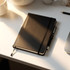 A black bound journal with an elastic closure and ribbon bookmark, placed on a desk beside a coffee cup and pen.