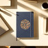 A navy soft-bound journal with a circular decorative design, placed on a desk beside a cup and stationery.