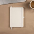 A white recycled A5 notebook with a ribbon bookmark, lying on a desk next to a laptop and a cup of coffee.