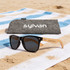 A pair of black sunglasses with wooden arms, alongside a grey pouch featuring a logo, on a sandy beach towel.