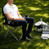 An oversized folding chair in black and silver with drinks in the cup holders, set on green grass. Accessories are nearby.