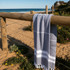 A grey and white Turkish towel draped over a wooden fence near a sandy beach and ocean.