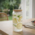 A glass tumbler with a bamboo lid and straw, filled with water, ice, lemon slices, and mint leaves.