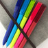 Five plastic pens in blue, green, yellow, pink, and red, arranged on a grey notebook and secured with an elastic band.