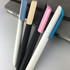Four plastic pens in white, black, light blue, and pink, arranged on a grey notebook. The pens feature different cap designs and a logo.
