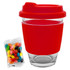 A glass coffee cup with a red silicone lid and sleeve, accompanied by a pack of colourful jelly beans.
