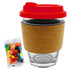 A glass coffee cup with a cork band and a red lid, accompanied by a pack of colourful jelly beans.