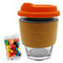 A glass coffee cup with an orange lid and cork band, accompanied by a bag of colourful jelly beans.