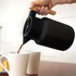 A drink bottle in black with a wooden handle, featuring a pouring spout, being used to serve coffee into white mugs.