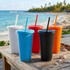A collection of plastic tumblers in blue, white, red, orange, and black with straws, set against a beach backdrop.