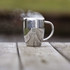 A silver coffee mug with a reflective surface and steam rising, placed on a wooden table. It features a logo.