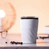 A drink mug in white with a twist design, featuring a black lid and an accompanying assortment of coffee beans.