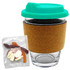A glass coffee cup with a cork band and a teal lid, accompanied by a pack of mixed lollies in various colours.