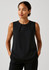 A sleeveless black blouse featuring pleat details, modelled by a woman with her hands in pockets.