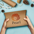 A person holding a large, brown pillow box with a logo on it, surrounded by crafting materials and pinecones.